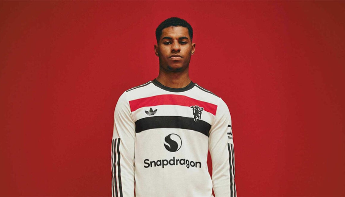 nova terceira camisa do Manchester United para a temporada 2024/2025. A camisa possui um design moderno e elegante, com base na cor off-white e detalhes em preto e vermelho nas faixas horizontais que representam a bandeira tricolor dos torcedores. A gola é redonda e em tom de carvão, enquanto o escudo do clube e o logo da Adidas aparecem em vermelho. 