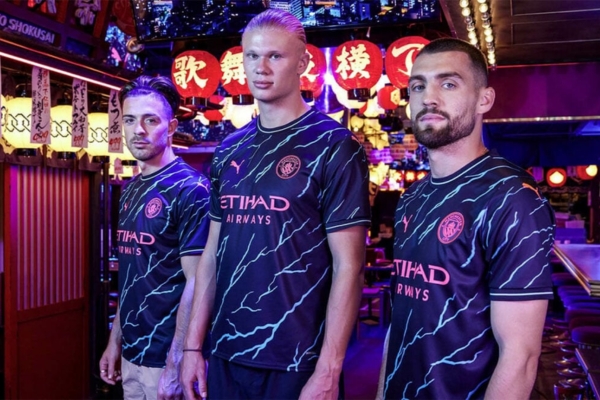 Terceira camisa azul marinho do Manchester City Puma 2023/24 com raios em azul celeste, escudo rosa, gola redonda e debrums nas laterais. Detalhes únicos representam a eletricidade do jogo e o estilo inigualável do time.