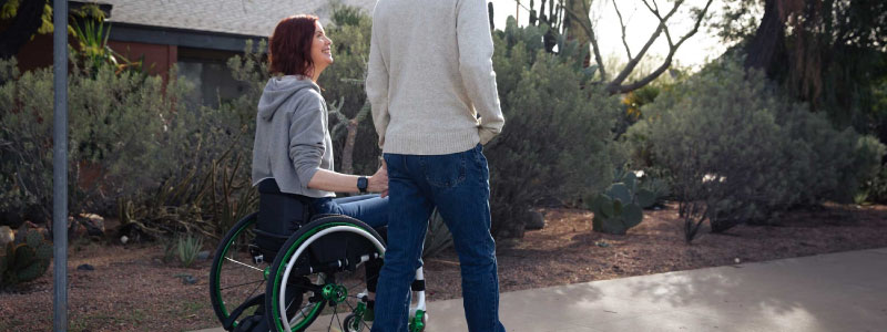 Mujer feliz en silla de ruedas acompañada por un hombre de espaldas, tomándole la mano en un día soleado. El cojín ROHO® MOSAIC® proporciona soporte y protección para prevenir úlceras por presión.