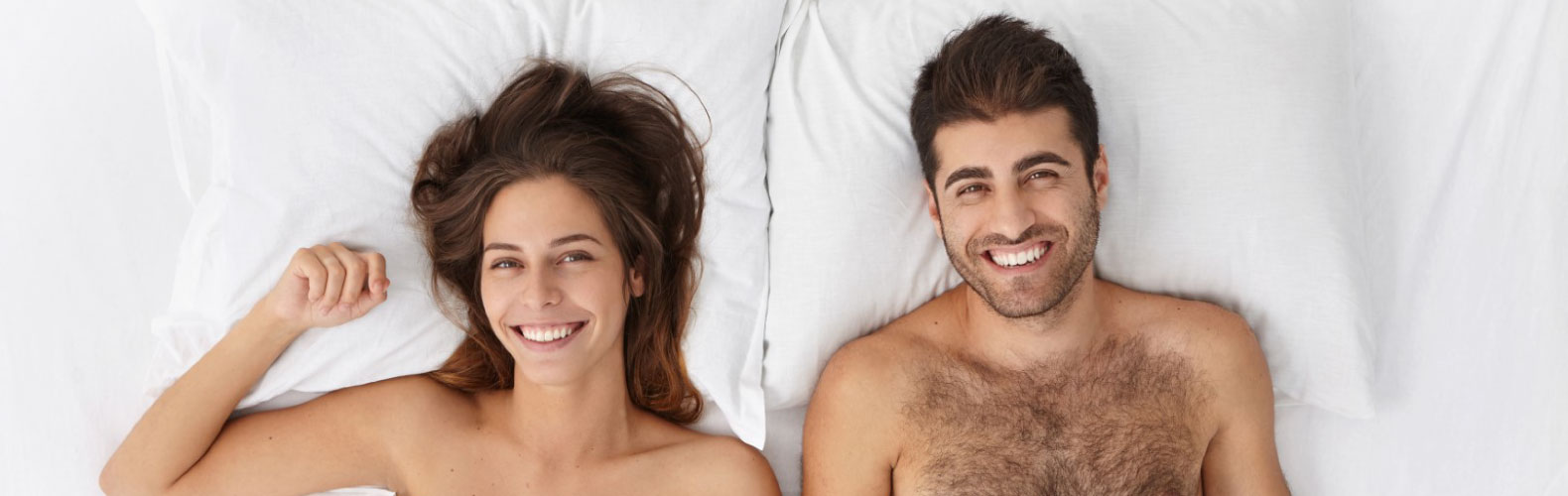 Una pareja sonriente y relajada se encuentra acostada en su cama, mirando hacia la cámara, mientras disfrutan de un momento de descanso y comodidad juntos.