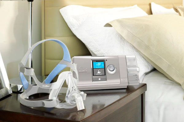 Banner del AirCurve™ 10 vAuto CPAP Binivel (vPAP) con una mascarilla oronasal f20 sobre un buró junto a la cama