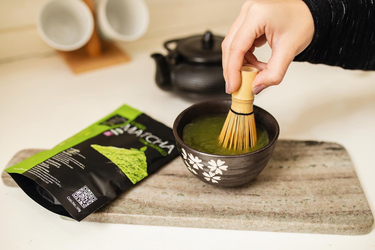 Bolsa de té matcha Kumori Tea junto a una taza. Una mano bate el té con un chasen, creando una mezcla suave y aromática. Disfruta de la experiencia única de calidad premium con cada sorbo.