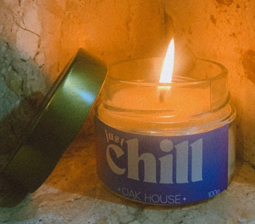 Imagem de uma vela acesa, ao lado dela tem uma tampa dourada e no pote de vidro tem a logo escrito Just Chill - Oak House