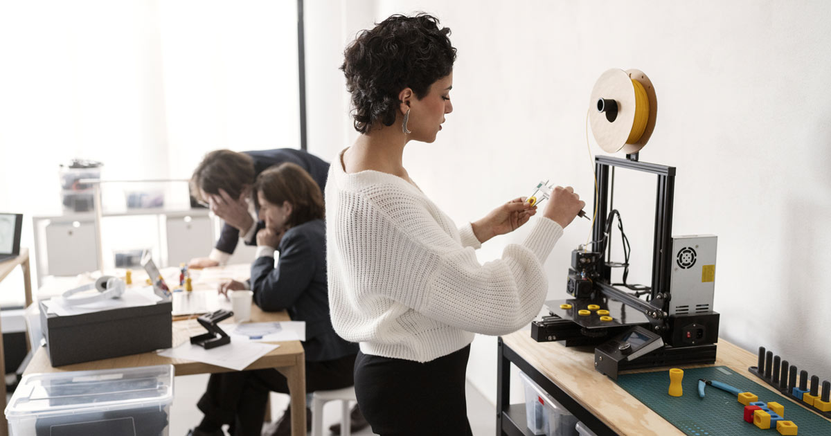 sala de escritório com três pessoas: uma mulher em primeiro plano medindo peças próximo à uma impressora 3D, e ao fundo um homem e uma mulher usam um computador