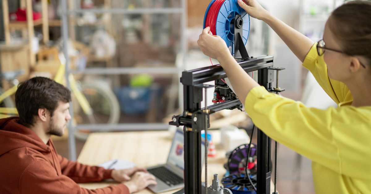 mulher ajustando um filamento em impressora 3D, ao fundo um homem usa um computador