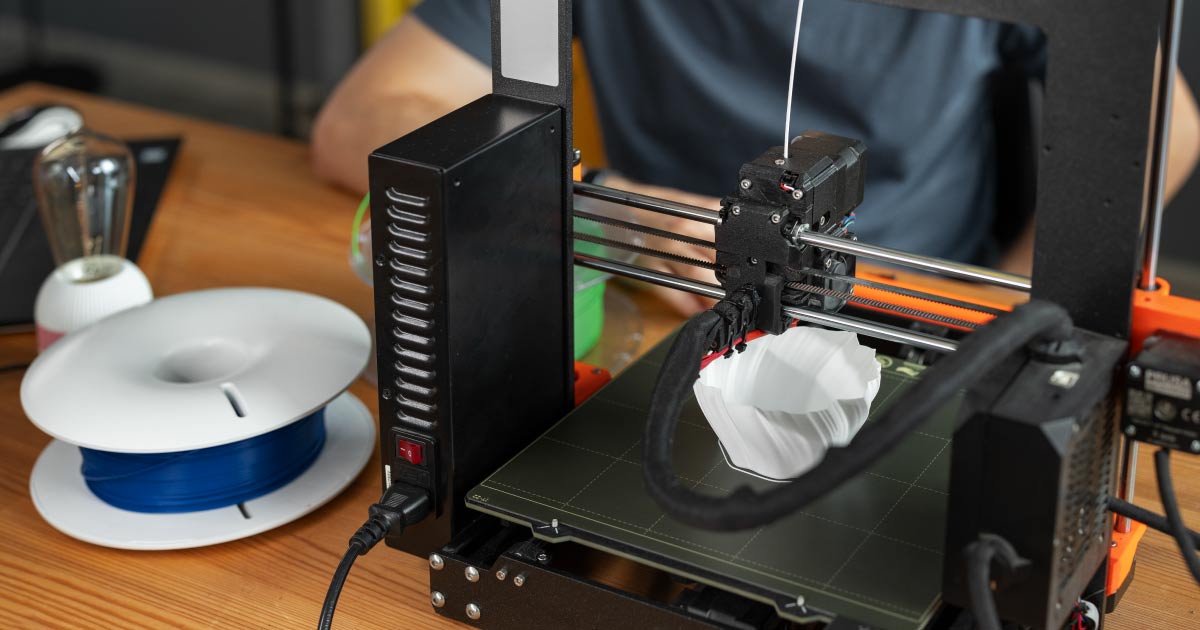 detalhe de uma impressora 3D aberta imprimindo uma peça, ao lado está um carretel de filamento 3D