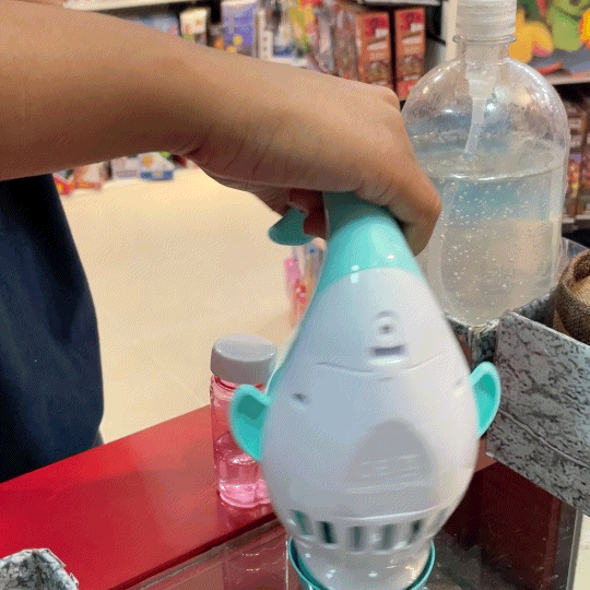 Uma mão segurando um brinquedo de tubarão azul e branco, enquanto faz bolhas de sabão. No fundo, é possível ver uma garrafa de plástico e outras embalagens de brinquedos.